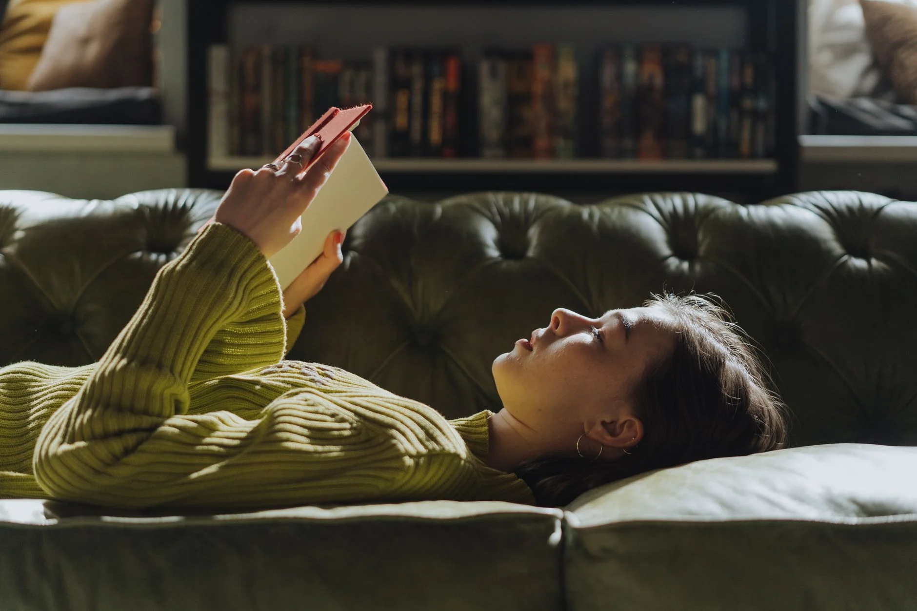 A young woman lying on a green sofa reading a book