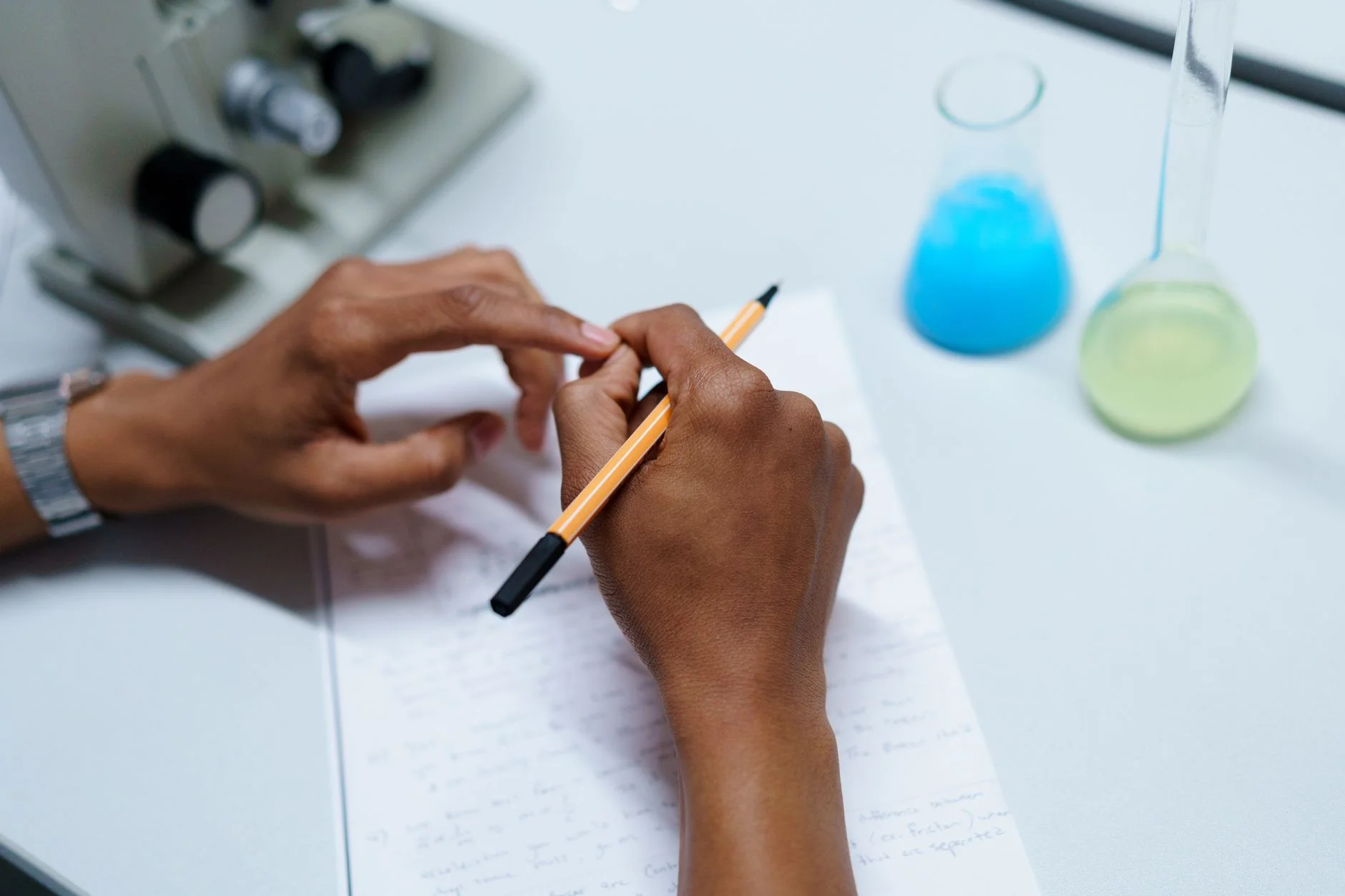 Hands writing notes on paper beside a microscope and two flasks of blue and green liquid in a laboratory