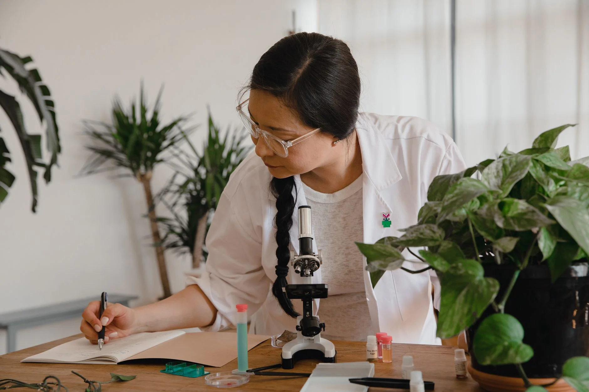 A woman in a white lab coat writing notes next to a microscope and a leafy plant