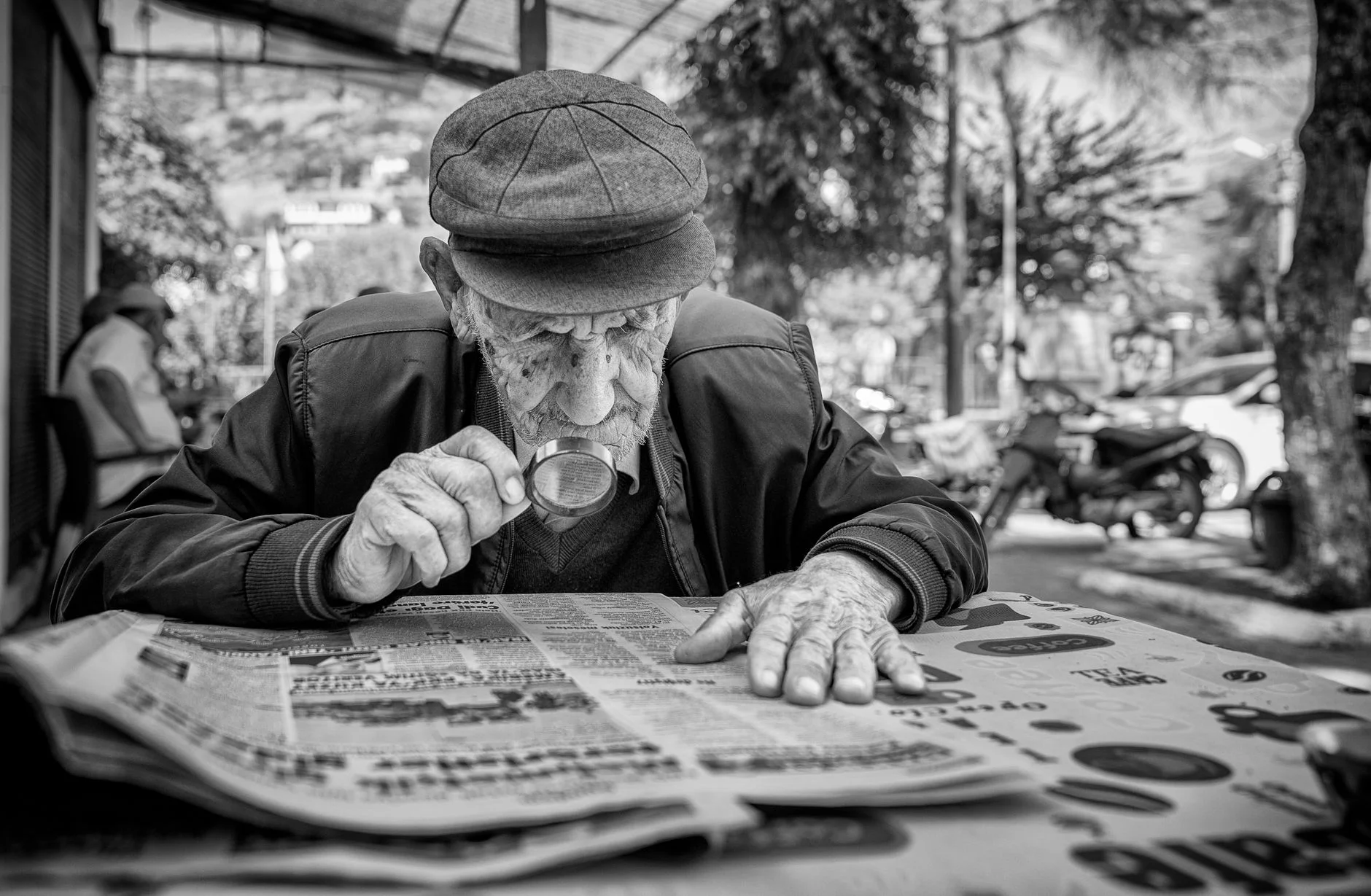 A man in a flat cap inspecting a newspaper closely through a small magnifying glass