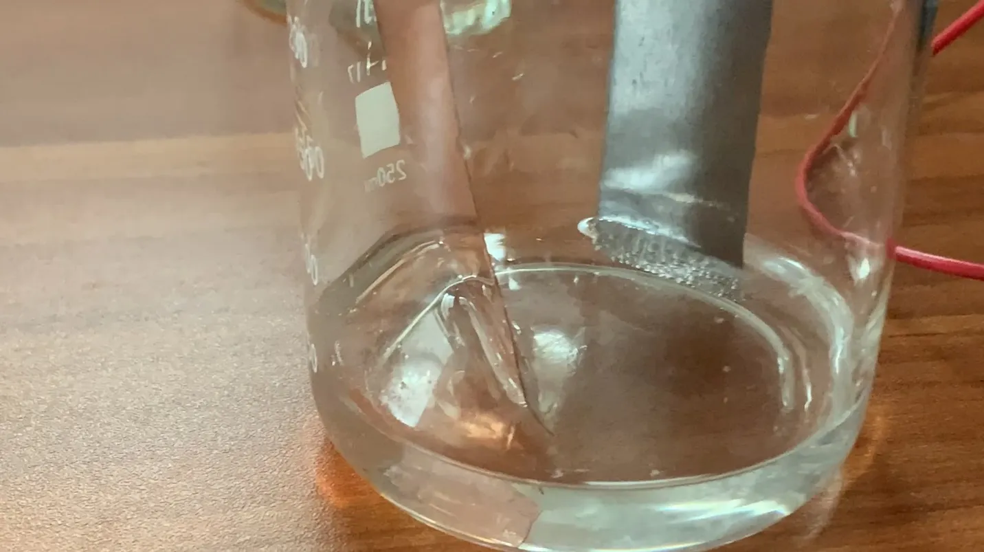 Close-up of a beaker of dilute sulfuric acid with a copper strip on the left and a grey zinc strip on the right, a line of small gas bubbles clinging to the zinc at the acid surface