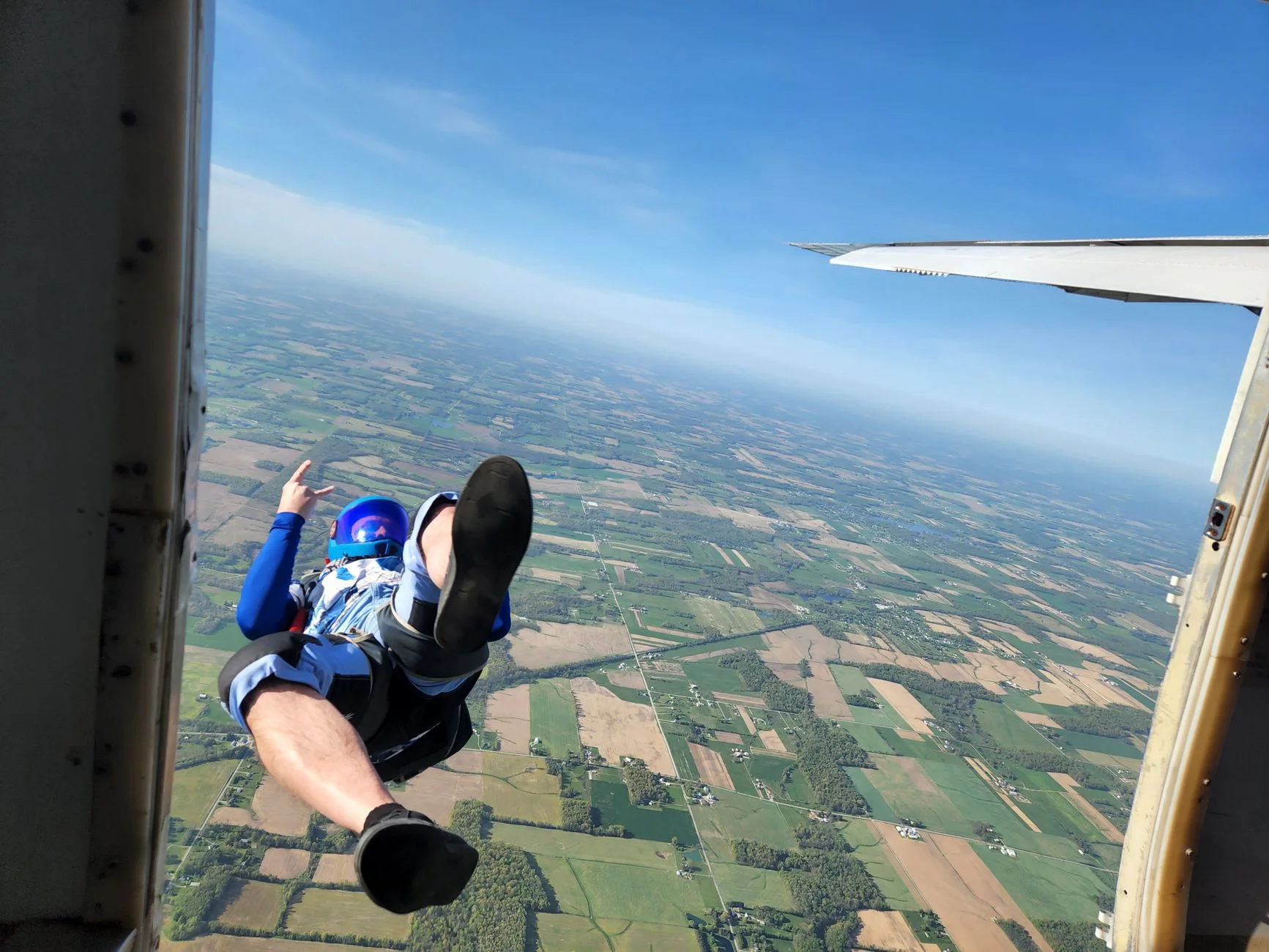 A skydiver leaping out of an aircraft door high above patchwork farmland