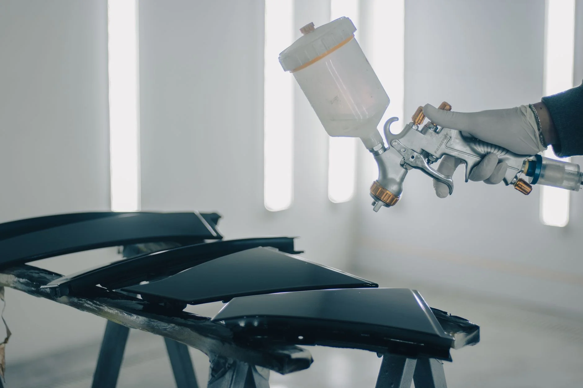 A gloved hand holding a spray gun over car panels in a paint booth