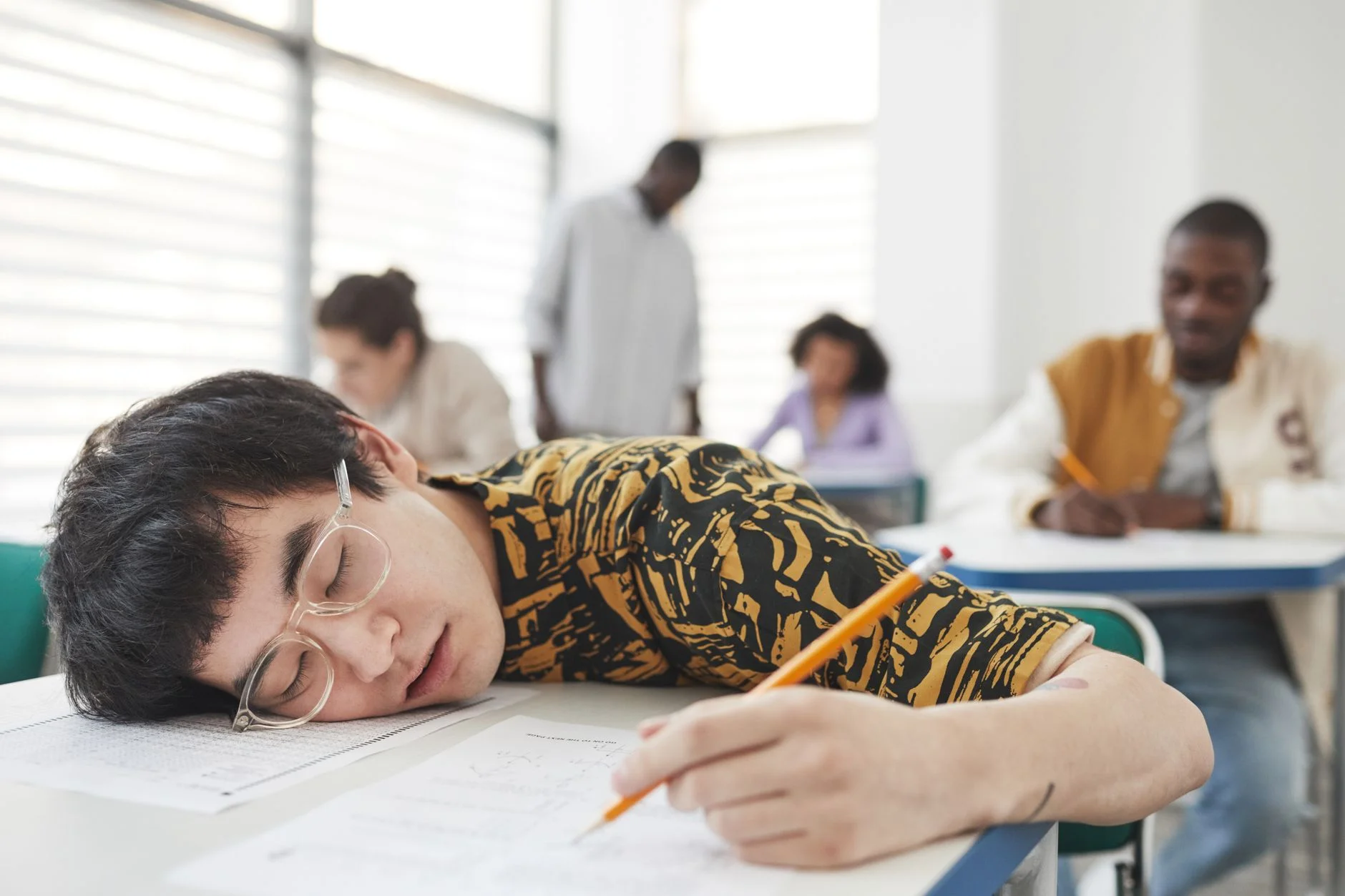 A student asleep with his head on an exam paper in a classroom while other students write