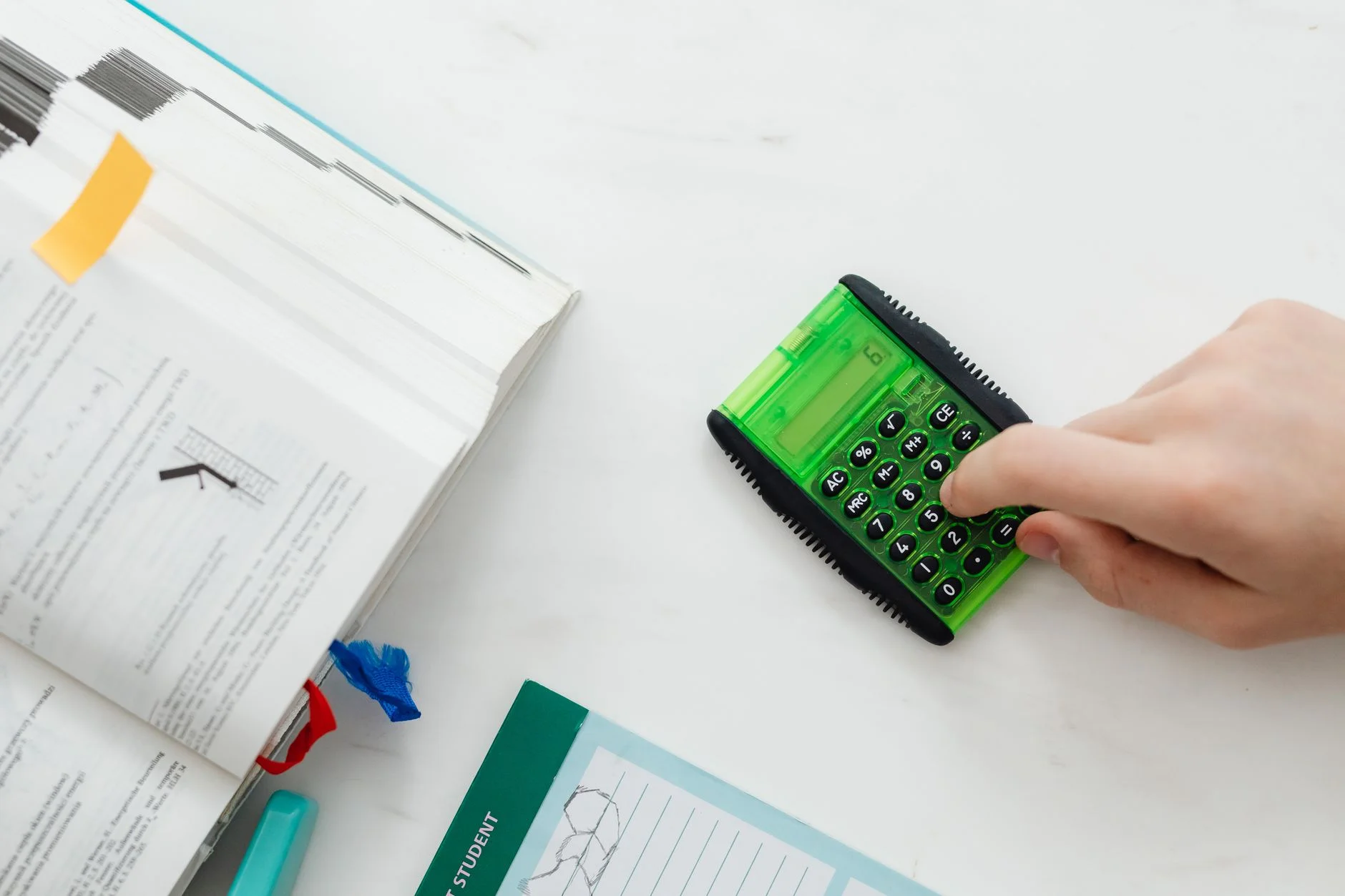 A hand pressing the buttons of a green calculator beside an open maths textbook