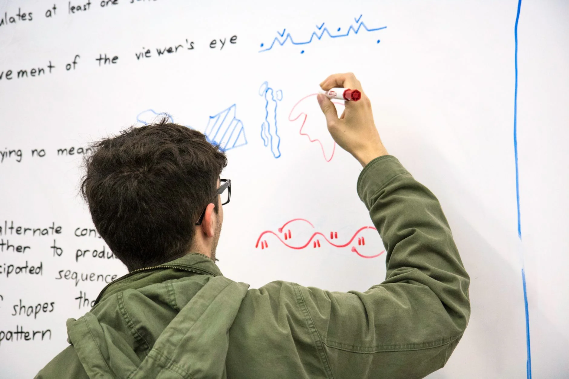 A student in a green jacket drawing colourful diagrams with a marker on a whiteboard covered in handwritten notes