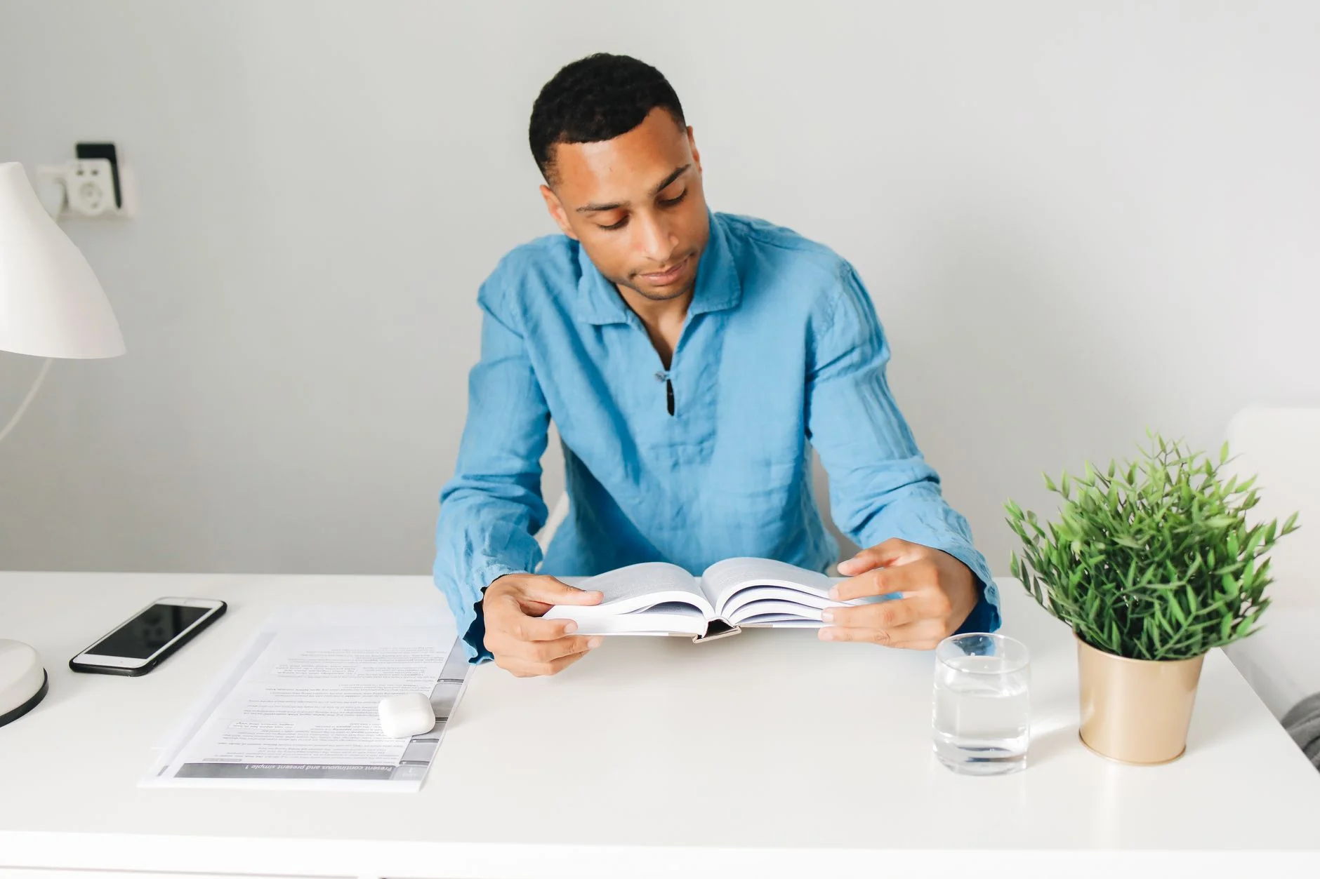 Young man reading a book at a tidy desk, with a phone lying on the desk beside him