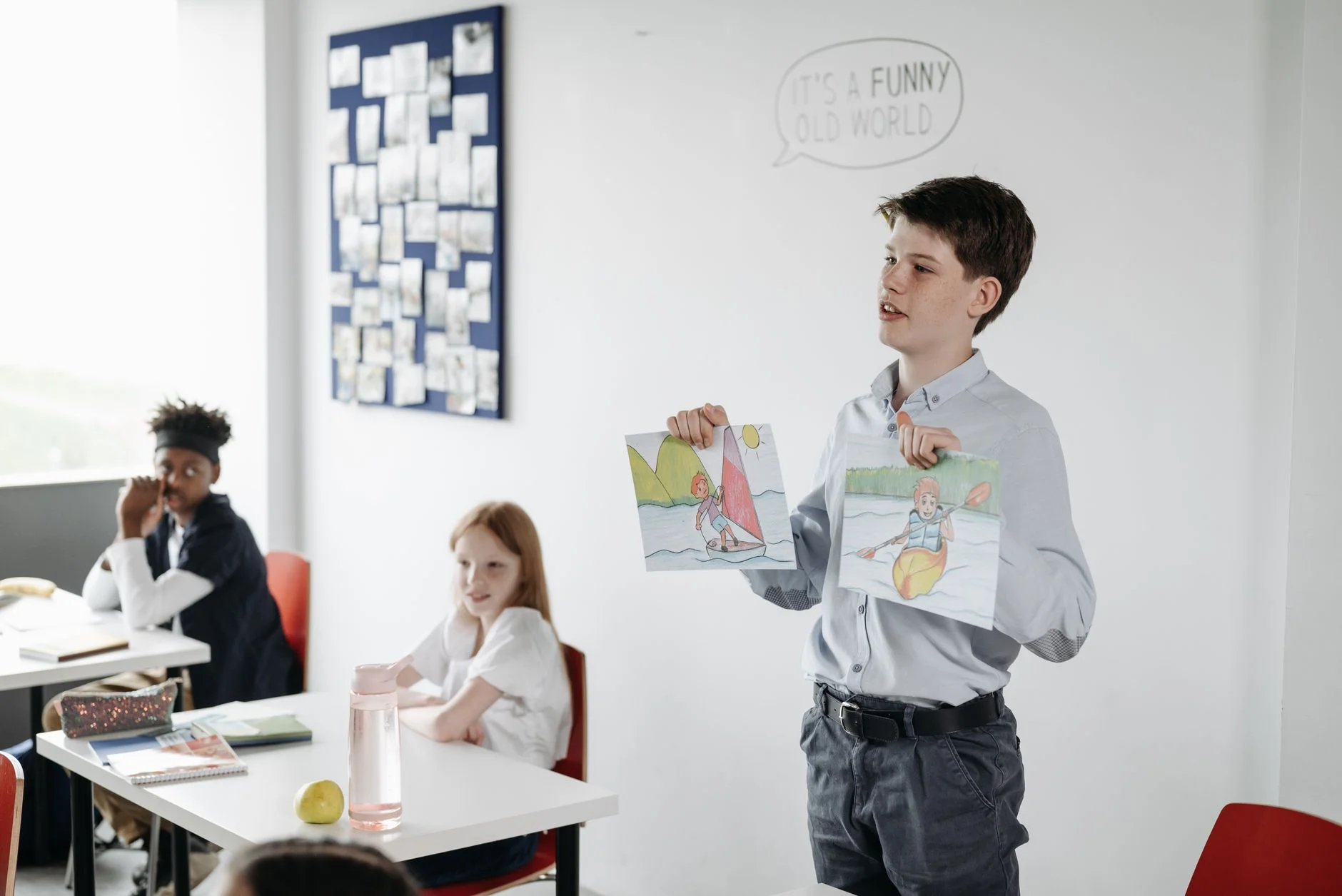 A student standing at the front of a bright classroom holding up two drawings while presenting to classmates