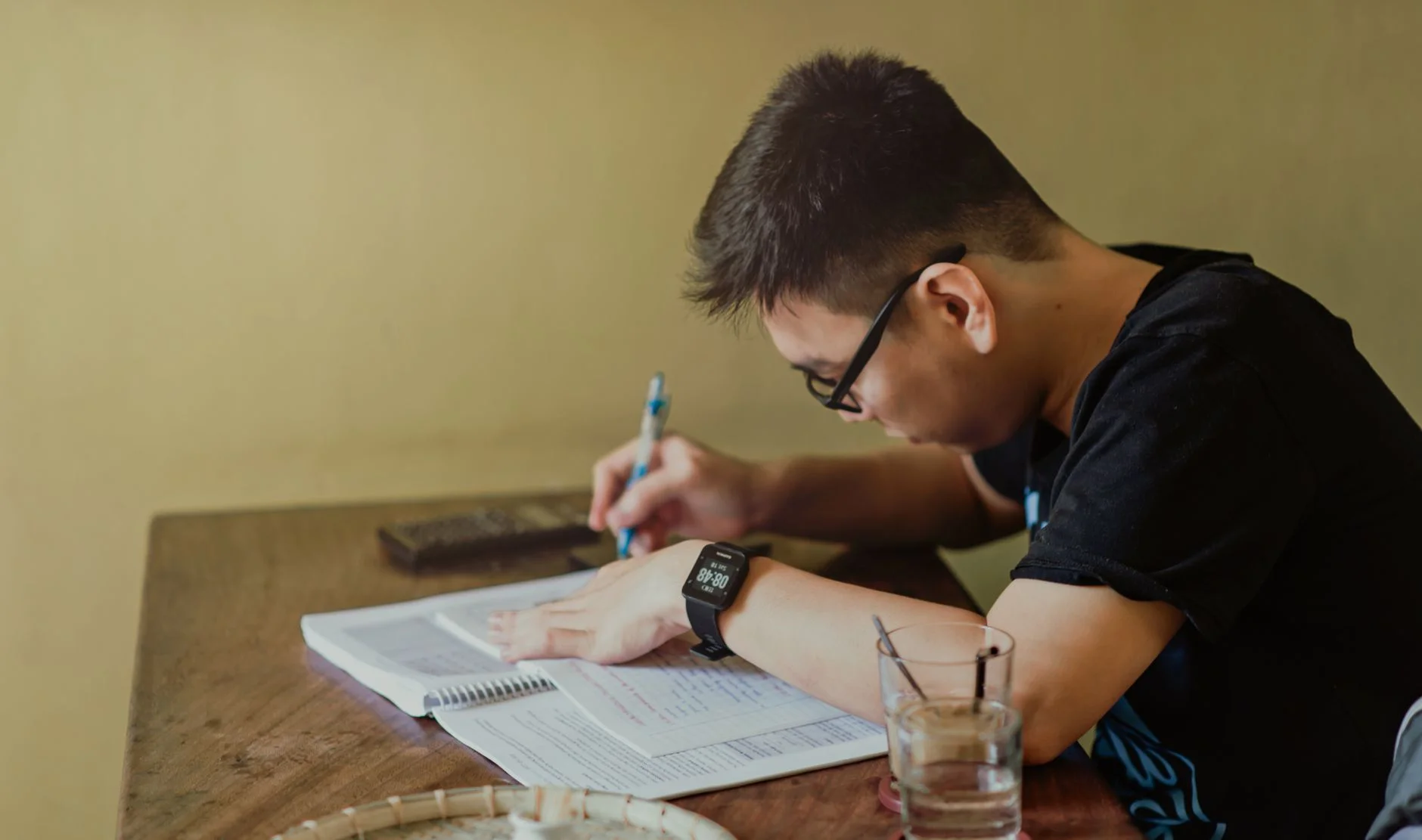 A student with glasses writing on a notepad beside an open book at a wooden desk