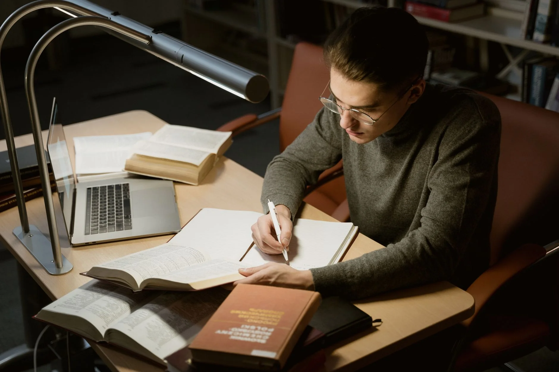 A student with glasses writing notes at a desk beside open books, a laptop and a lamp