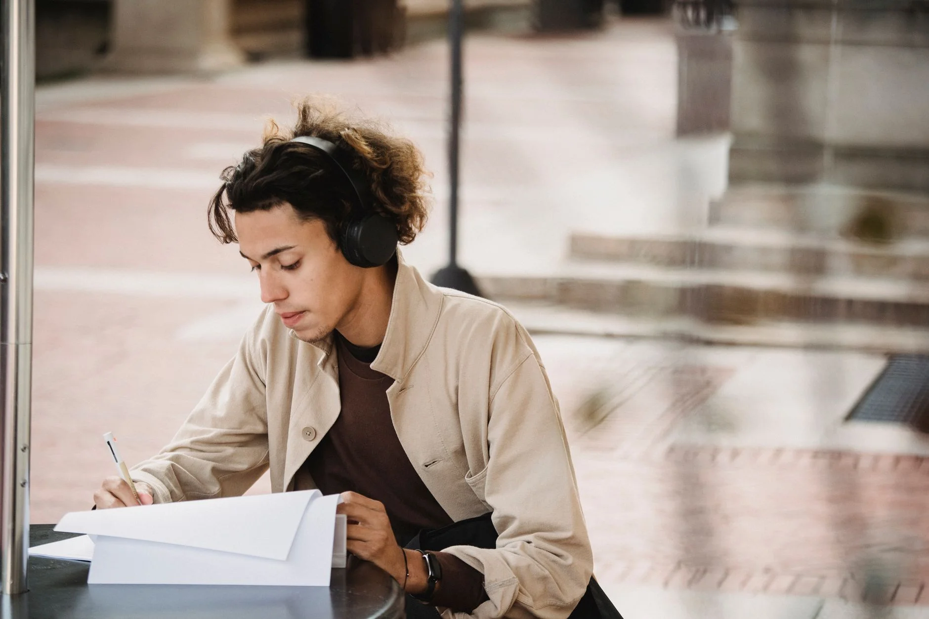 A young man wearing headphones writing notes on paper at an outdoor table