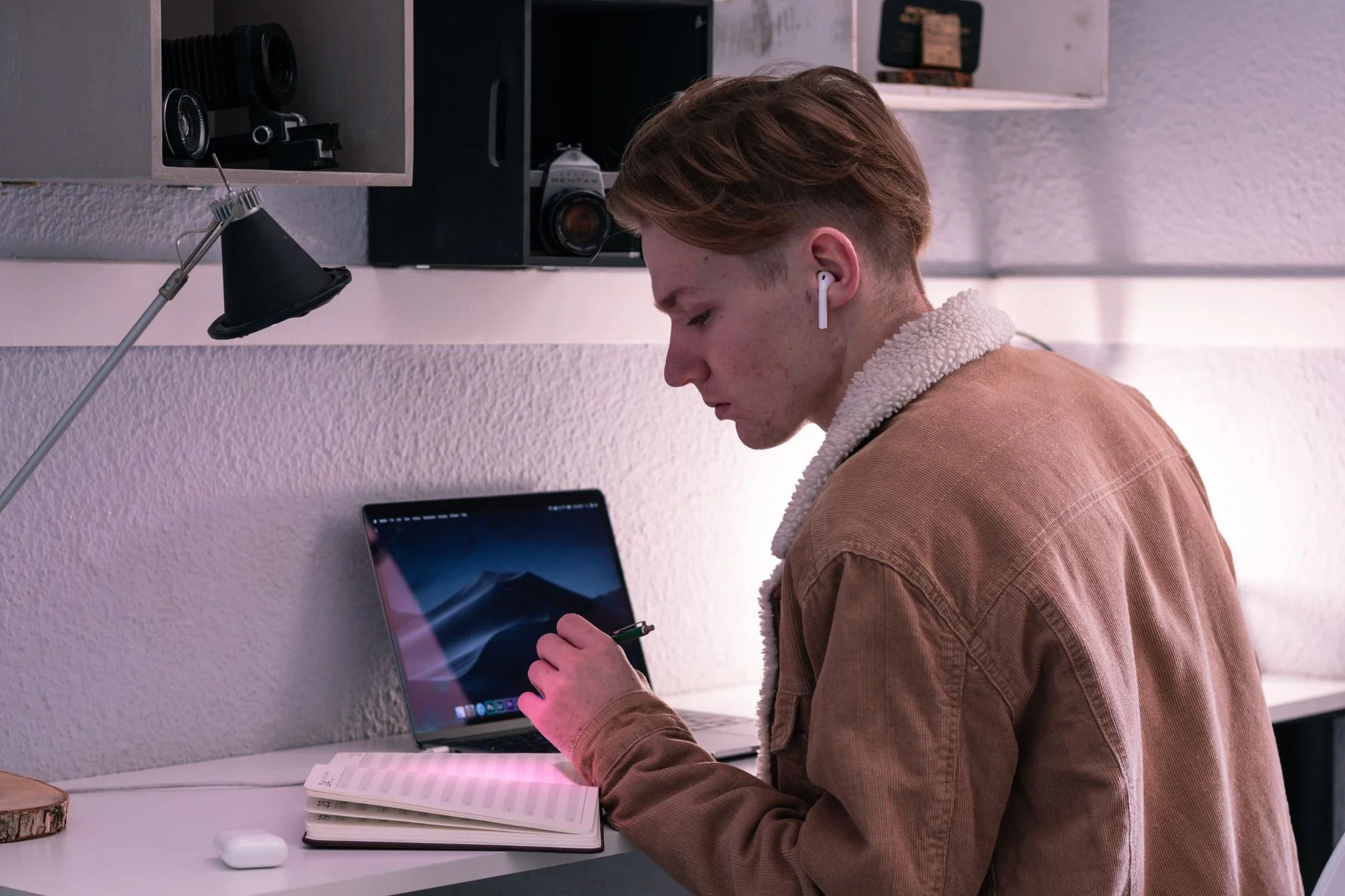 A teenage student wearing earbuds writing in a planner at a desk with a laptop