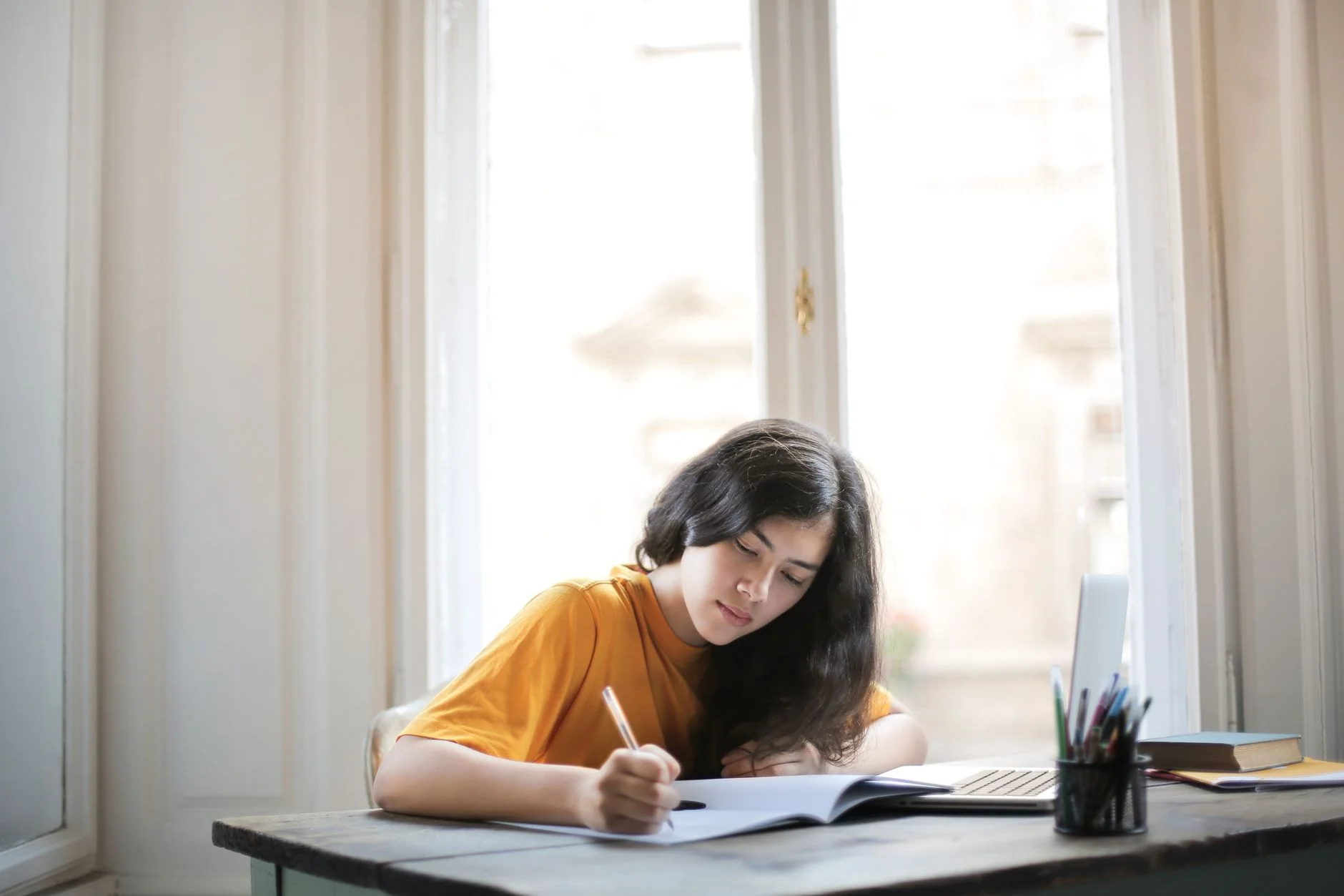 A student in a yellow top writing in a notebook at a wooden desk beside a window