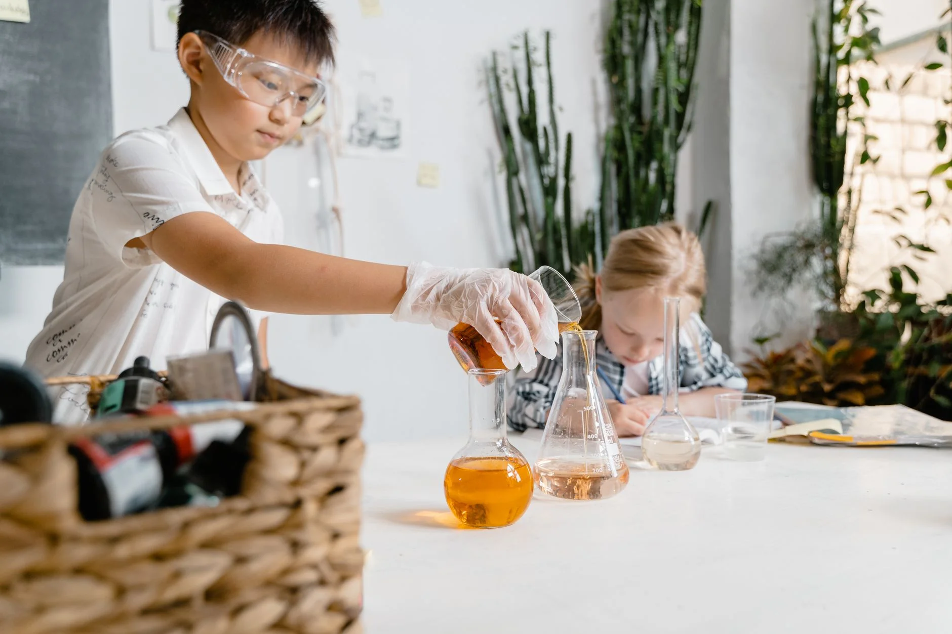 A child in safety goggles and a glove pouring orange liquid into a conical flask on a bench while another child writes behind