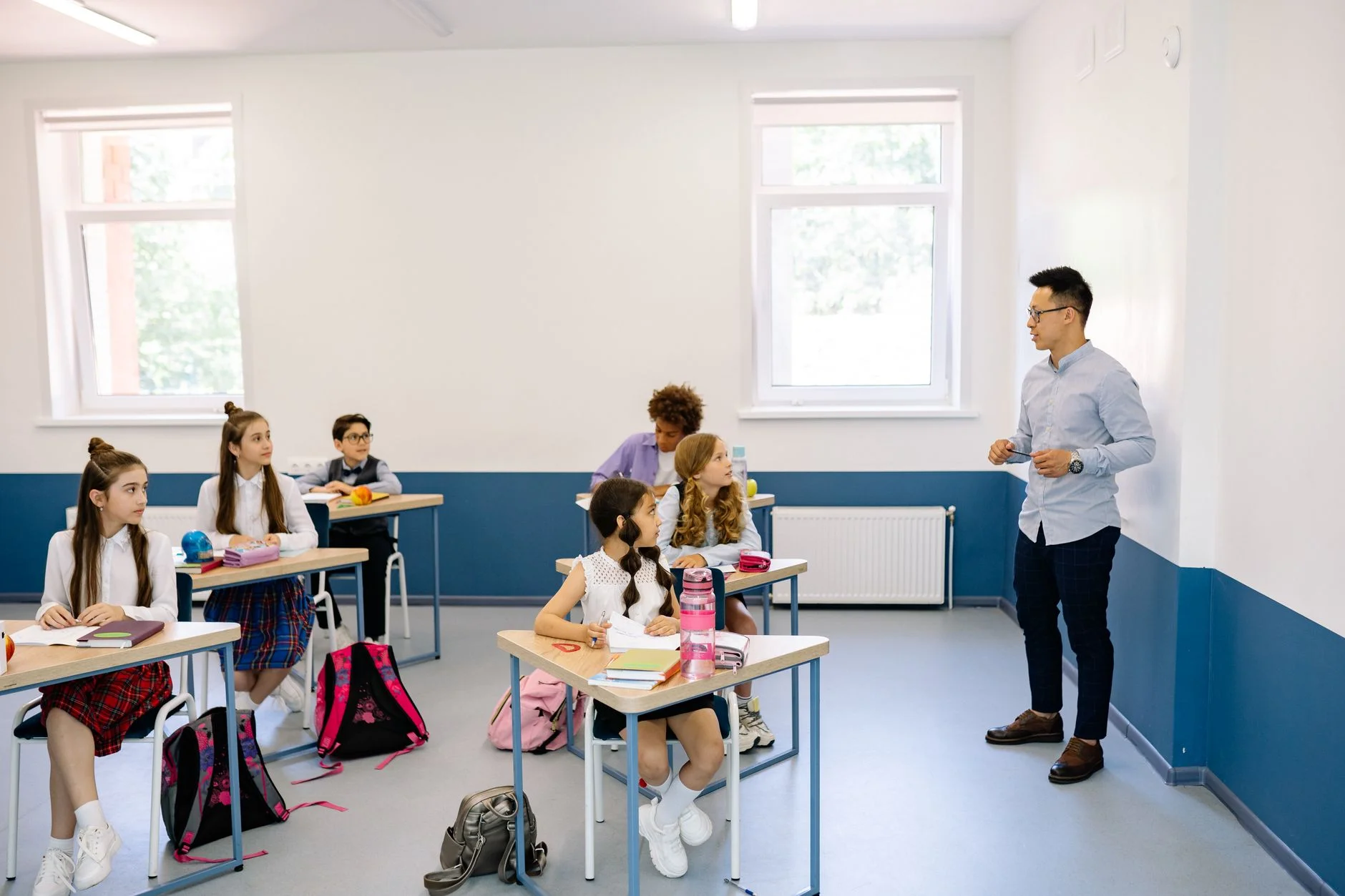 Pupils in school uniform at their desks turned towards a teacher who is explaining something at the front of a bright classroom