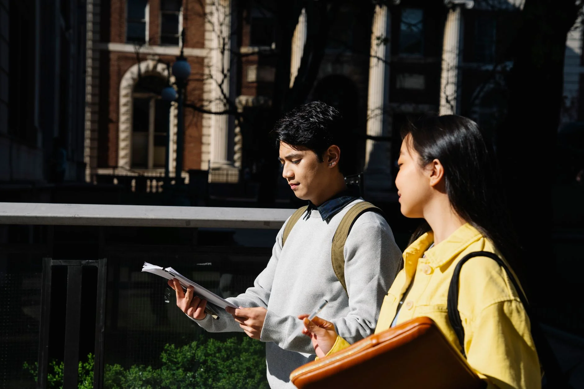 Two students walking outdoors on a campus, one holding loose papers and the other a folder, talking about their notes