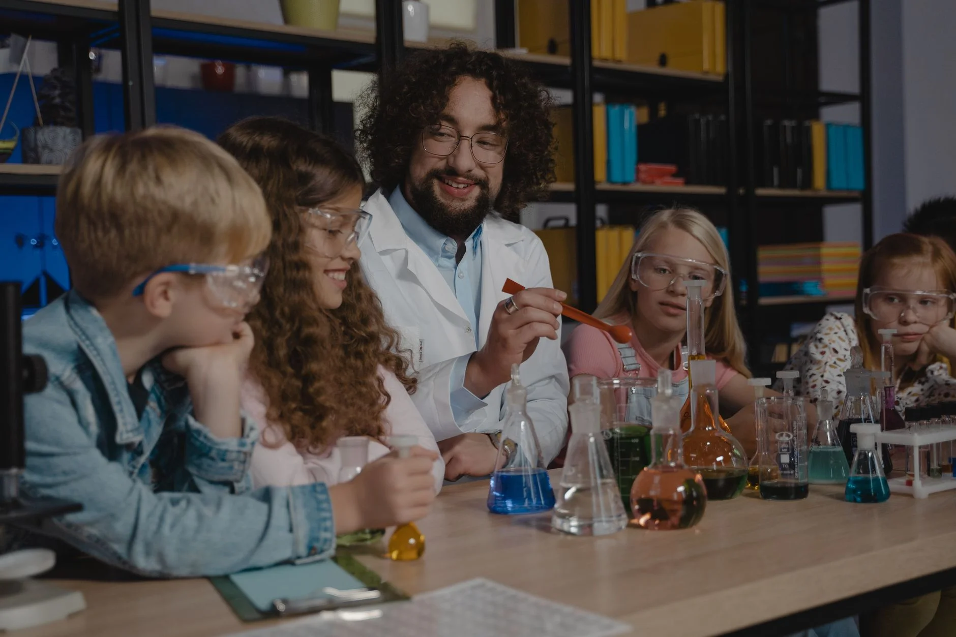 A group of students in safety goggles watching a teacher at a table of coloured liquids in flasks