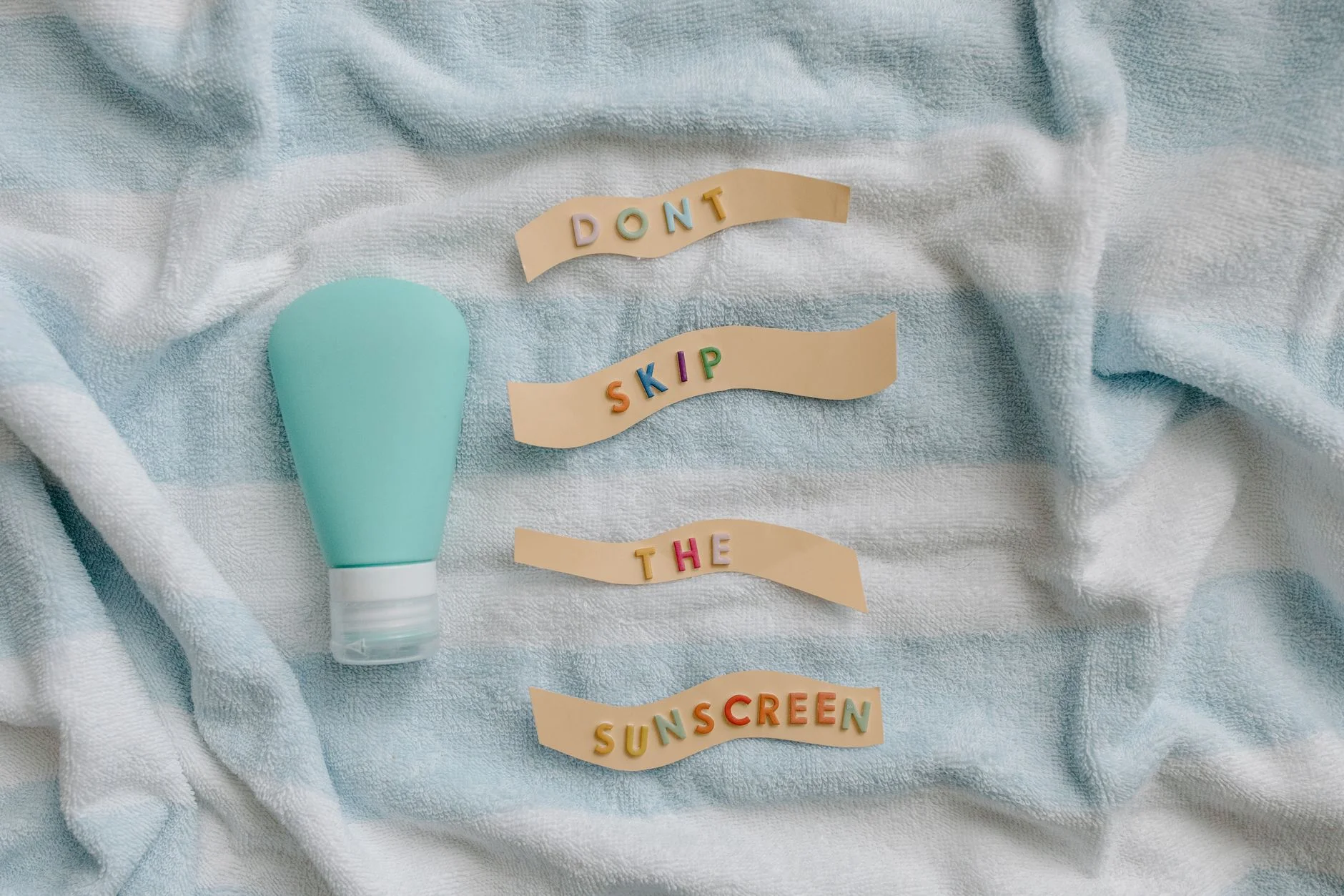 A pale turquoise (mint green) bottle on a striped towel next to paper strips spelling out “Don’t skip the sunscreen”