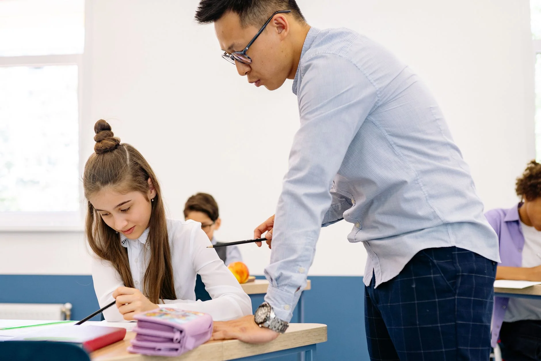 A teacher leaning over a desk pointing at a pupil’s work while she writes