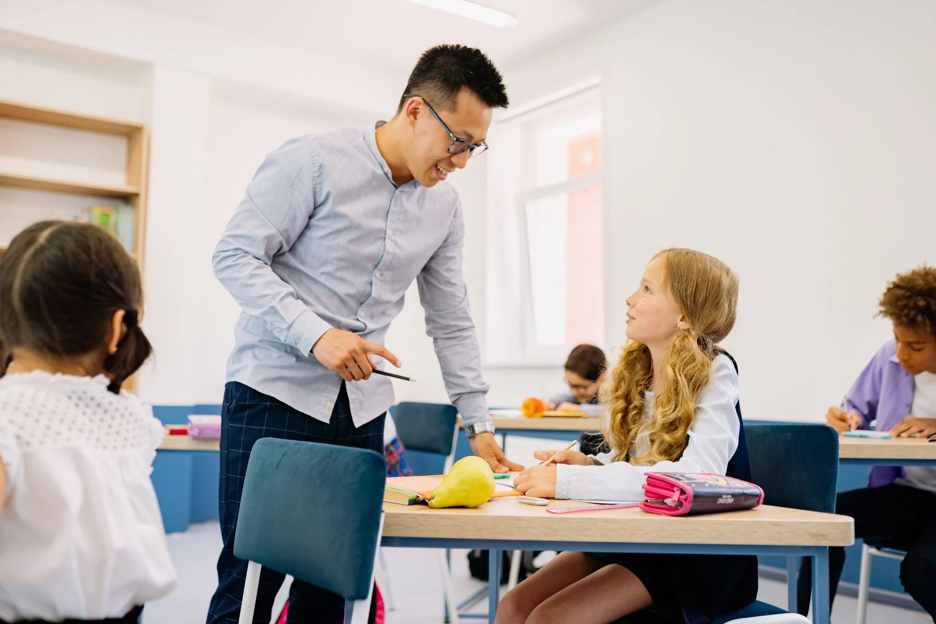 A teacher leaning over a desk and smiling while a pupil looks up at him