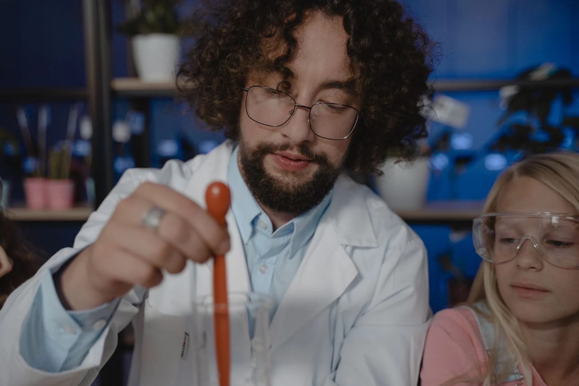 A teacher in a lab coat holding a glass pipette next to a student wearing safety goggles