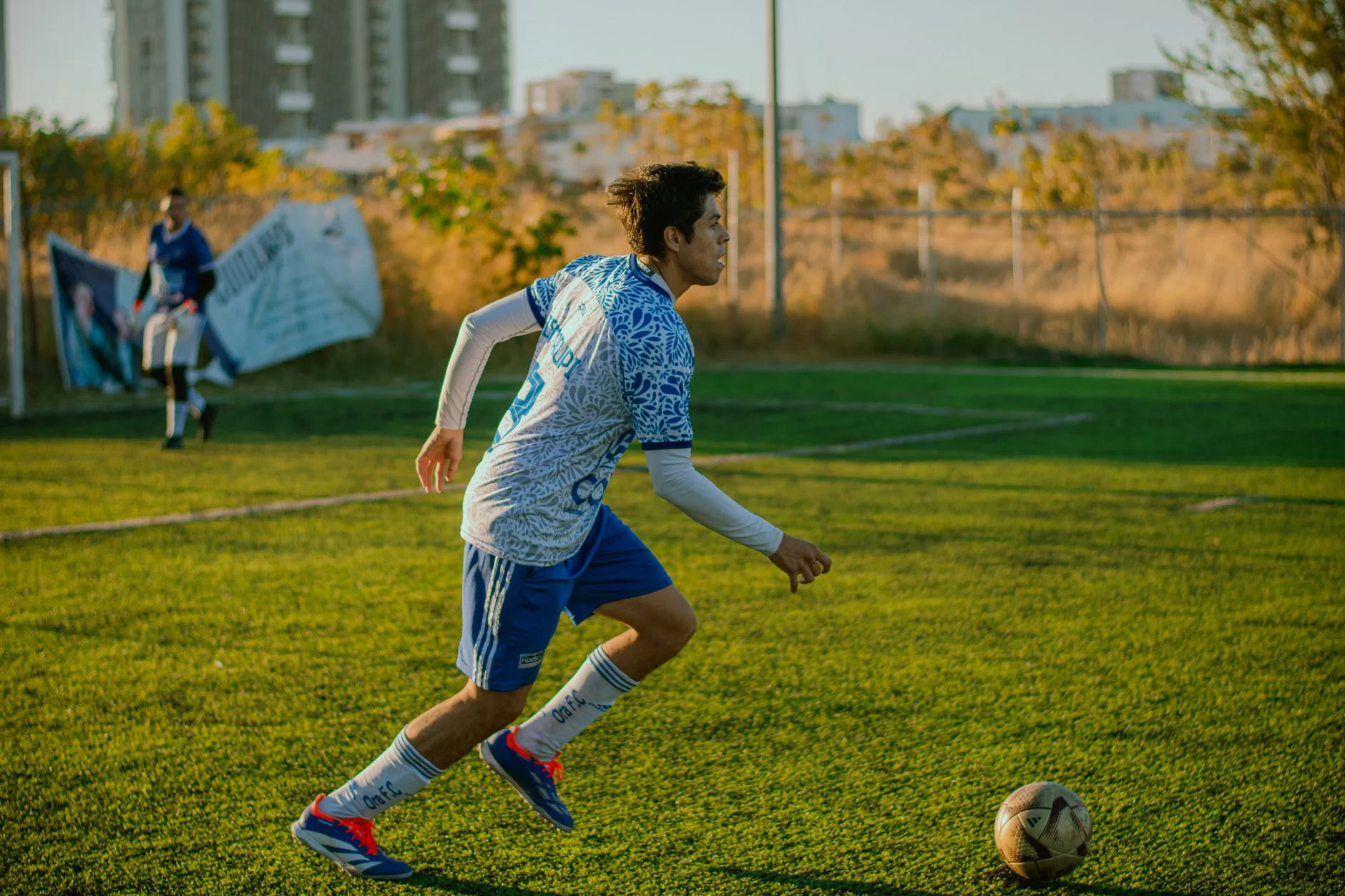 A teenage footballer in a blue kit dribbling a ball on a grass pitch