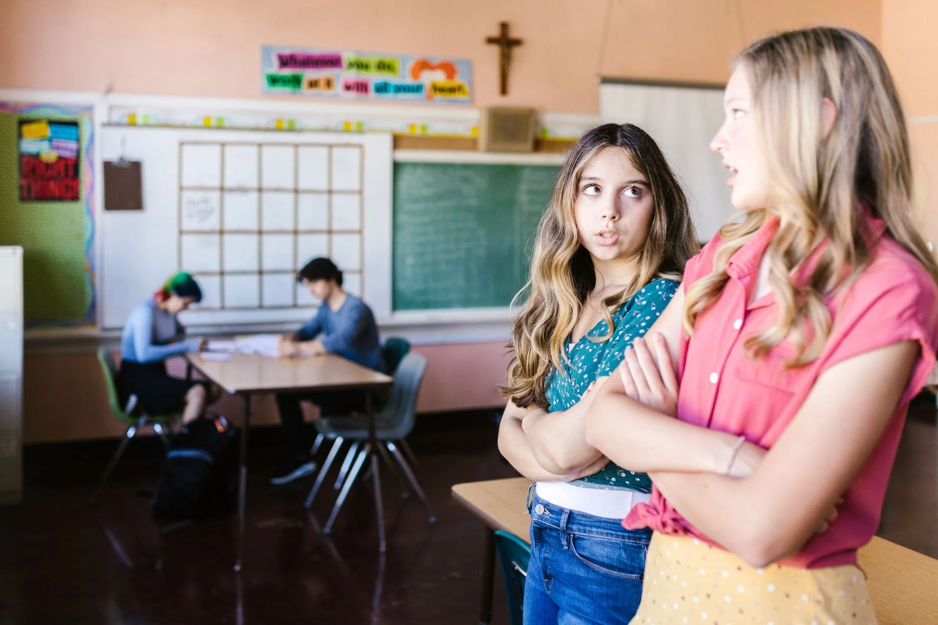 Two teenage girls standing with folded arms in a classroom, one talking while the other looks away
