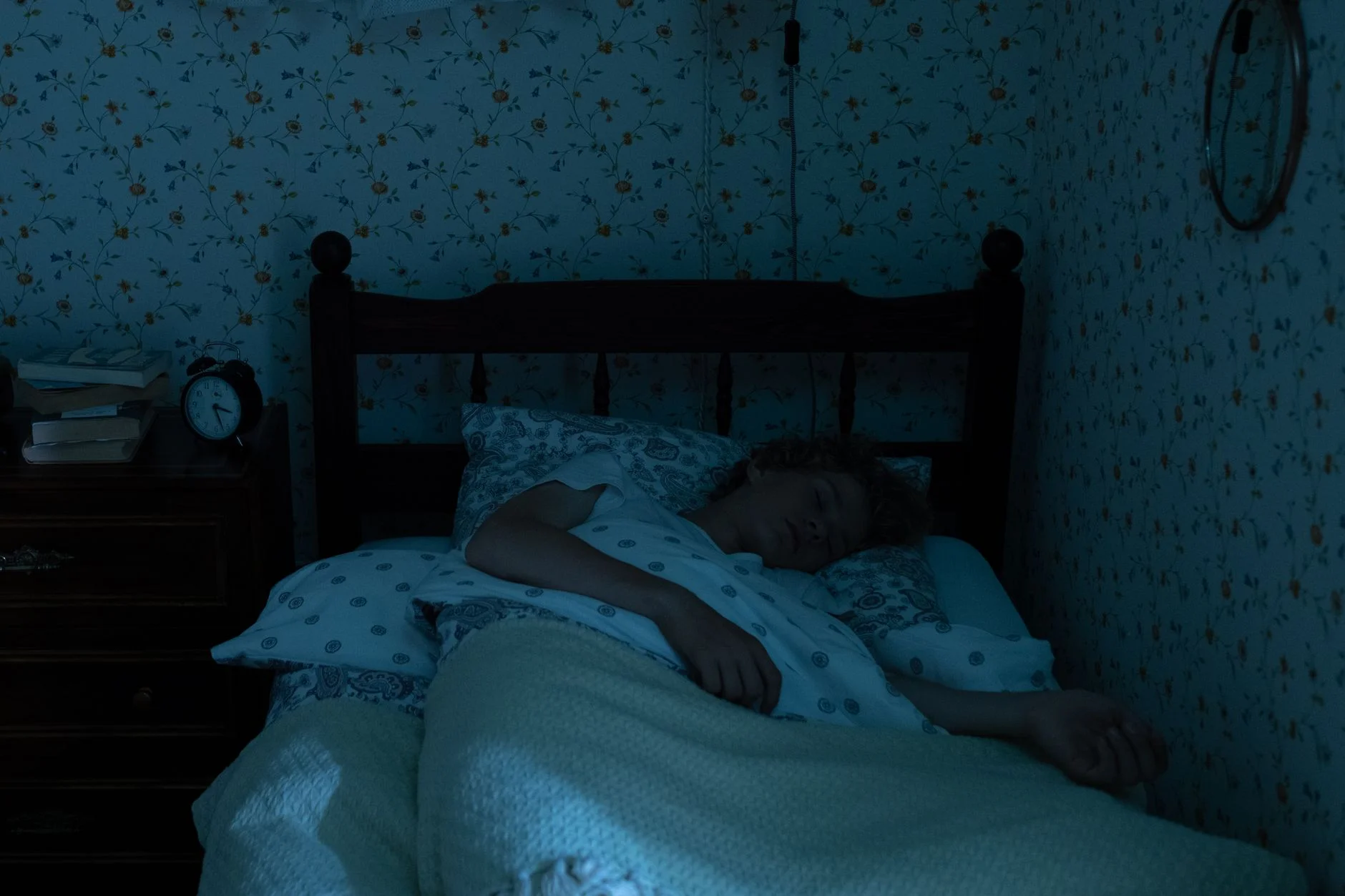 A person asleep in a dark bedroom with an alarm clock and books on the bedside table