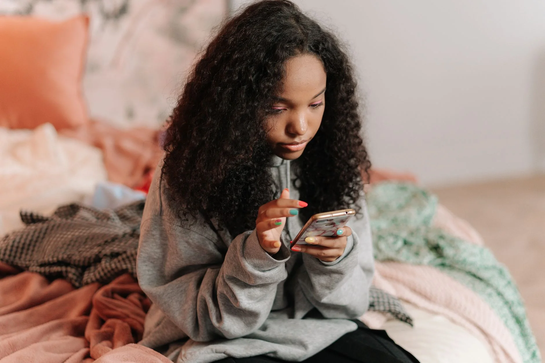A teenager with curly hair sitting on a bed looking at a smartphone