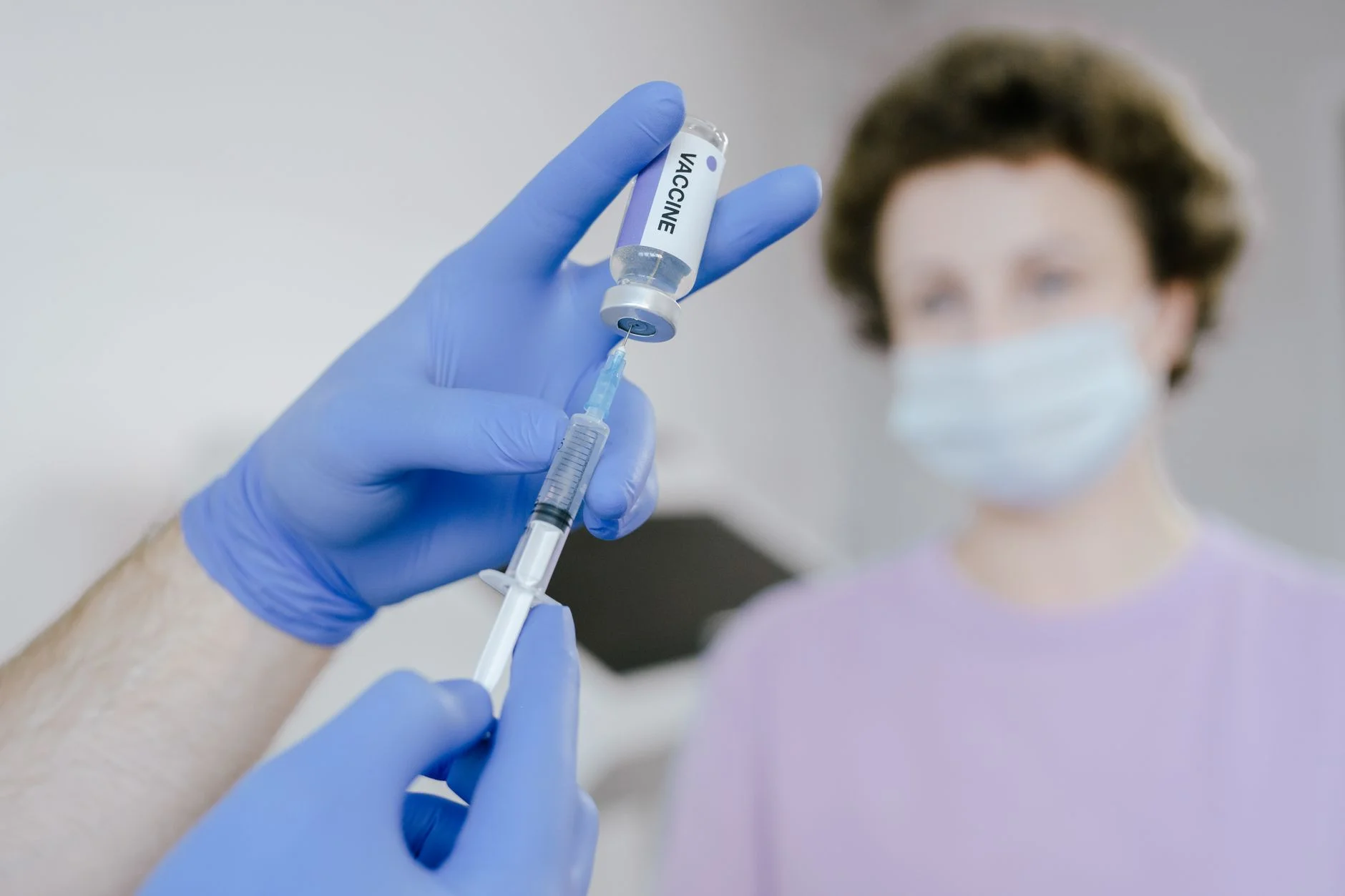 A health worker in blue gloves filling a syringe from a vaccine vial, with a patient behind