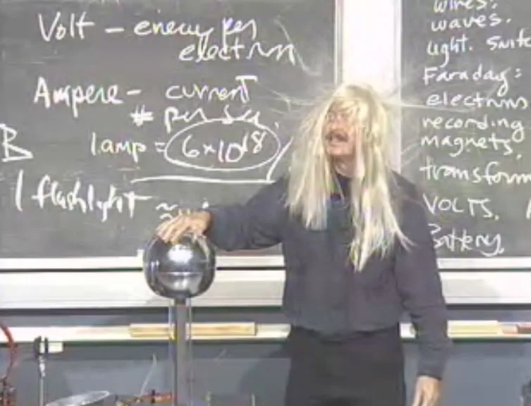 A physics lecturer with long hair standing on end while touching the dome of a Van de Graaff generator