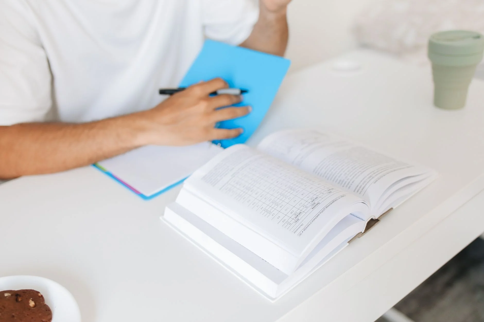 A person writing in a notebook beside an open book on a white desk